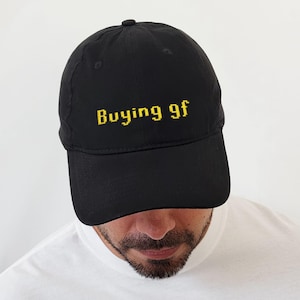 May include: Black baseball cap with the text "Buying gf" embroidered in yellow. The cap has a curved brim and a rounded crown. The hat is a classic style, suitable for casual wear.