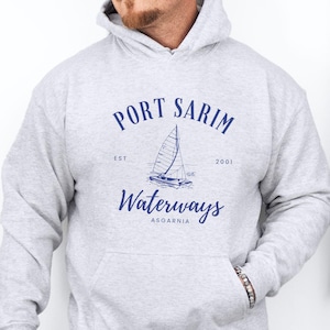 May include: A light gray hoodie with a front pocket and a drawstring hood. The navy blue graphic on the front reads "PORT SARIM WATERWAYS ASGARNIA" with a sailboat illustration. The hoodie is worn by a person.