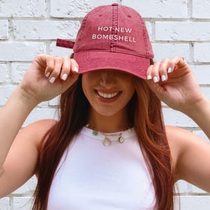 May include: A vintage red baseball cap featuring the text HOT NEW BOMBSHELL embroidered in white on the front. The cap has a curved brim, a structured crown, and an adjustable strap at the back. The fabric has a slightly faded, worn-in appearance.