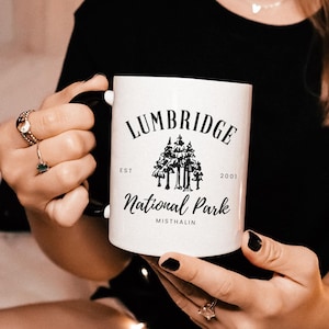 May include: White ceramic mug with a black design that says "Lumbridge National Park Est. 2001 Misthallin". The design features a group of trees.