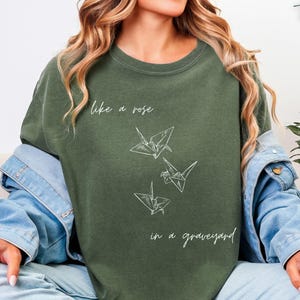 May include: Olive green t-shirt with the text "like a rose in a graveyard" and three origami cranes. The word "Hemp" is written in cursive in the upper right corner. The shirt is part of a casual clothing collection.