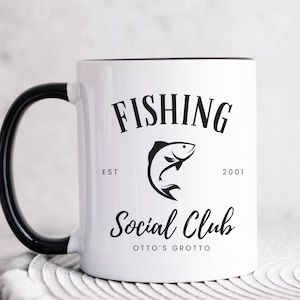 May include: White ceramic coffee mug with a black handle and black rim. The mug has a black and white graphic of a fish with the text "Fishing Social Club Est 2001 Otto's Grotto".
