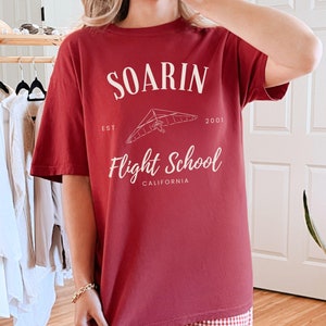 May include: A red t-shirt with a white graphic design that says "Soarin' Flight School Est. 2001 California" with a drawing of a hang glider.