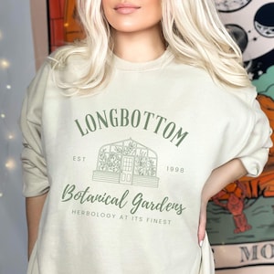 May include: A light beige sweatshirt with a green graphic design that says "Longbottom Botanical Gardens Est. 1998 Herbology at its finest". The graphic features a greenhouse with plants inside.