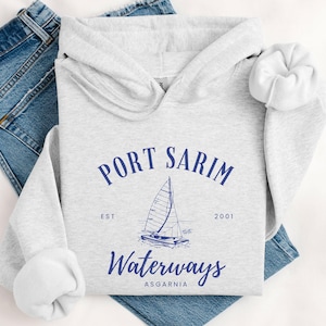 May include: A light gray hoodie with the text "PORT SARIM WATERWAYS ASGARNIA" in navy blue, along with a sailboat graphic. The hoodie is laid on top of a pair of blue jeans and a white scrunchie.
