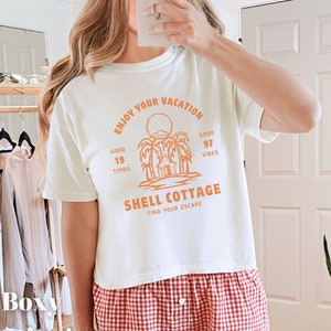 May include: White boxy t-shirt with an orange graphic design that says "Enjoy Your Vacation" with palm trees and a small house. The text also says "Good 19 Times", "Good 97 Vibes", "Shell Cottage", and "Find Your Escape".