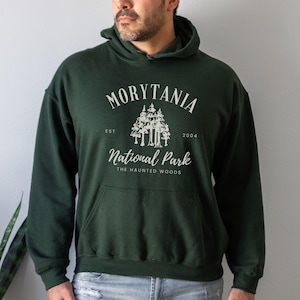 May include: Dark green hooded sweatshirt with a front pocket. The white graphic reads "Morytania National Park, The Haunted Woods" with a tree illustration. The year "2004" is also included in the design. The sweatshirt is made of a soft material.