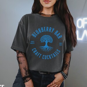 May include: Dark gray t-shirt with a blue graphic that reads "Blurberry Bar Craft Cocktails" around a tree design. The shirt has a relaxed fit and short sleeves. The text "Open Late" is visible in the background.