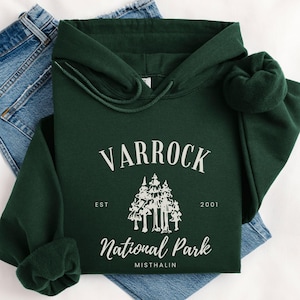 May include: A dark green hoodie with the text "VARROCK National Park MISTHALIN" in white. The hoodie has a drawstring hood and long sleeves. The hoodie is folded on top of a pair of blue jeans.