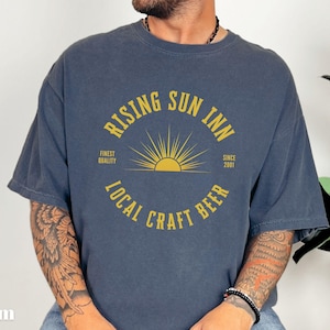 May include: Denim blue t-shirt with a distressed look. The shirt features a yellow graphic with the text "RISING SUN INN" and "LOCAL CRAFT BEER". The design includes a sunburst and the words "Finest Quality" and "Since 2001".