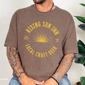 May include: Espresso-colored t-shirt with a circular graphic that reads "Rising Sun Inn Local Craft Beer" in yellow. The design includes a sunburst and the text "Finest Quality Since 2001".