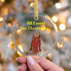 May include: Clear glass Christmas ornament with gold ribbon. The ornament features the text "All I want for Christmas..." in yellow pixelated font, and a red and gold cape design.