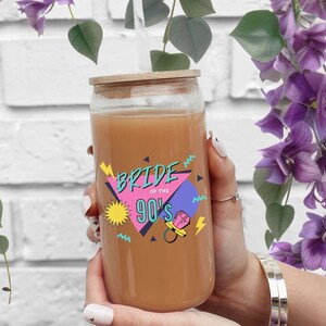 May include: A clear glass can with a wooden lid and a straw. The can has a pink, blue, yellow, and black graphic that says "Bride of the 90's" with a pacifier and a ring. The can is filled with a light brown liquid.