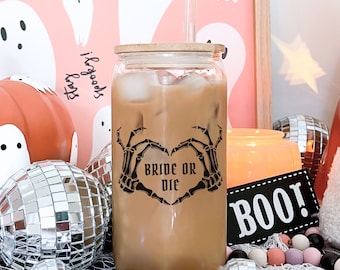 May include: A clear glass with a bamboo lid and a black skeleton hand design that says "Bride or Die" filled with iced coffee. The glass is surrounded by Halloween decorations, including ghosts, pumpkins, and disco balls.