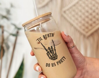 May include: A clear glass reusable iced coffee cup with a wooden lid and a straw. The cup has a black graphic of a skeleton hand giving the rock and roll sign with the text "'til death do us party".