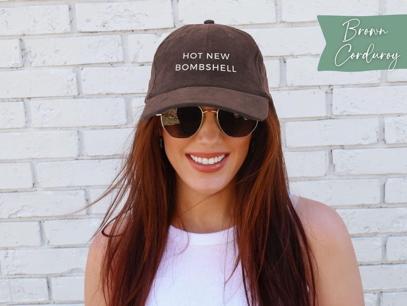 May include: A brown corduroy baseball cap featuring the text HOT NEW BOMBSHELL embroidered in white on the front panel. The cap has a structured crown, a curved brim, and a button on top. The fabric has a visible ribbed texture characteristic of corduroy.