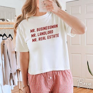 May include: Cream-colored t-shirt with the words "MR. BUSINESSMAN, MR. LANDLORD, MR. REAL ESTATE" in red. The person is wearing red and white checkered pajama pants. The image is taken indoors.