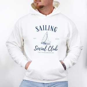 May include: Cream-colored hoodie with a front pocket and a drawstring hood. The navy blue graphic reads "SAILING SOCIAL CLUB" with a sailboat illustration. The text "EST 2025" and "THE PANDEMONIUM" are also included. A denim jacket hangs in the background.