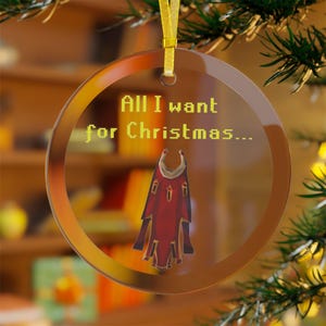 May include: Clear glass Christmas ornament with a gold ribbon. The ornament has a brown border and features the text "All I want for Christmas..." in yellow, along with a red and gold cape design.