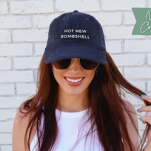 May include: A navy blue corduroy baseball cap featuring the text HOT NEW BOMSHELL embroidered in white on the front. The cap has a structured crown, a curved brim, and a button on top. The fabric has a visible ribbed texture characteristic of corduroy.