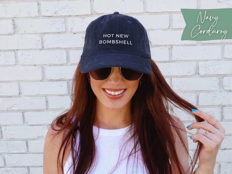 May include: A navy blue corduroy baseball cap featuring the text HOT NEW BOMSHELL embroidered in white on the front. The cap has a structured crown, a curved brim, and a button on top. The fabric has a visible ribbed texture characteristic of corduroy.