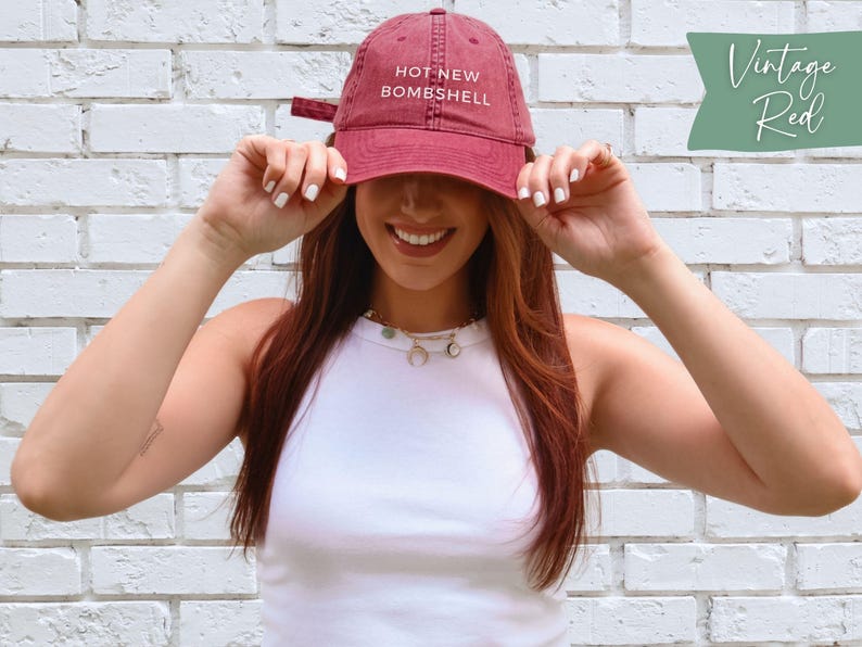 May include: A vintage red baseball cap featuring the text HOT NEW BOMBSHELL embroidered in white on the front. The cap has a curved brim, a structured crown, and an adjustable strap at the back. The fabric has a slightly faded, worn-in appearance.