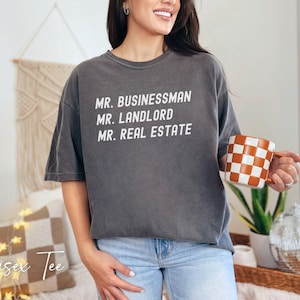Puede incluir: Camiseta unisex gris oscuro con las palabras "MR. BUSINESSMAN, MR. LANDLORD, MR. REAL ESTATE" en blanco. La persona sostiene una taza a cuadros naranja y blanca. Las palabras "Unisex Tee" est&aacute;n escritas en cursiva.
