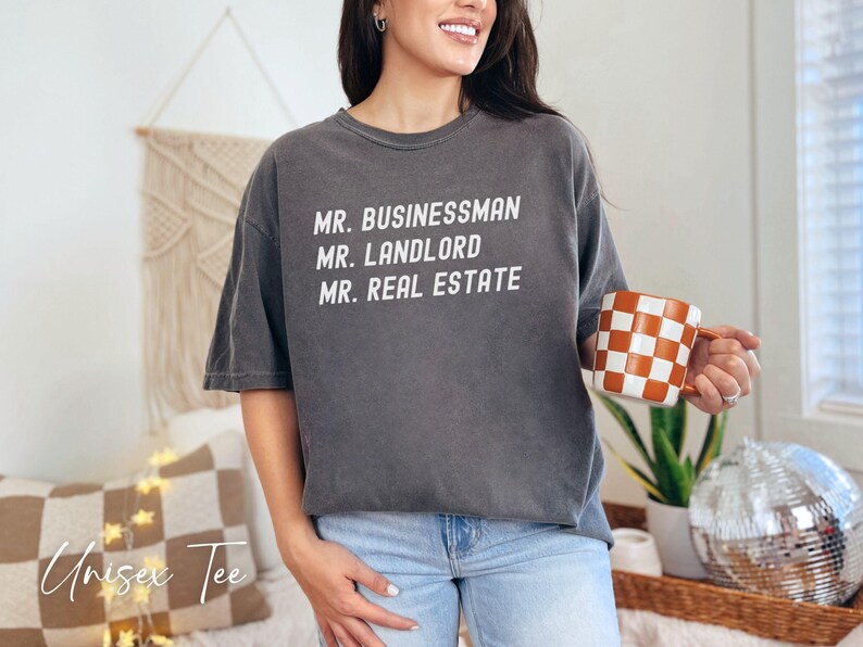 Puede incluir: Camiseta unisex gris oscuro con las palabras "MR. BUSINESSMAN, MR. LANDLORD, MR. REAL ESTATE" en blanco. La persona sostiene una taza a cuadros naranja y blanca. Las palabras "Unisex Tee" est&aacute;n escritas en cursiva.