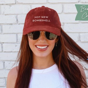 May include: A burgundy corduroy baseball cap featuring the text HOT NEW BOMBSHELL embroidered in white on the front. The cap has a structured crown, a curved brim, and small ventilation eyelets on the top panels of the fabric.