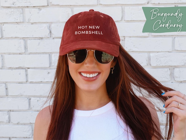 May include: A burgundy corduroy baseball cap featuring the text HOT NEW BOMBSHELL embroidered in white on the front. The cap has a structured crown, a curved brim, and small ventilation eyelets on the top panels of the fabric.