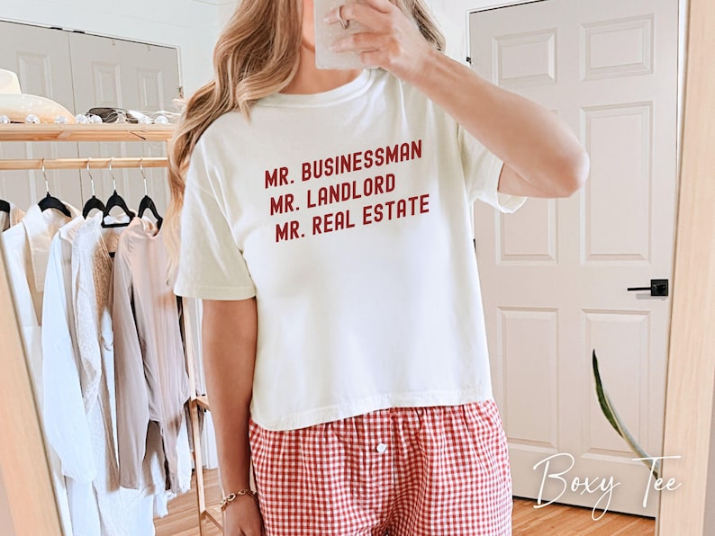 May include: Cream-colored t-shirt with the words "MR. BUSINESSMAN, MR. LANDLORD, MR. REAL ESTATE" in red. The person is wearing red and white checkered pajama pants. The image is taken indoors.