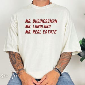 Puede incluir: Camiseta unisex color crema con las frases "MR. BUSINESSMAN", "MR. LANDLORD" y "MR. REAL ESTATE" impresas en rojo. La camiseta tiene mangas cortas y un corte holgado. Las palabras "Unisex Tee" est&aacute;n escritas en cursiva en la parte inferior.