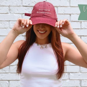 May include: A vintage red baseball cap featuring the text HOT NEW BOMBSHELL embroidered in white on the front. The cap has a curved brim, a structured crown, and an adjustable strap at the back. The fabric has a slightly faded, worn-in appearance.