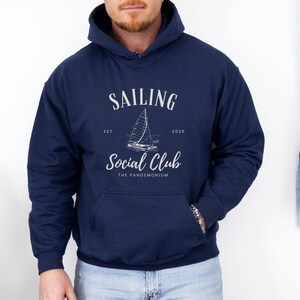 May include: Navy blue hooded sweatshirt with a white graphic that reads "SAILING Social Club" with a sailboat illustration. The text "EST 2025" is also included. The sweatshirt has a front pocket and a drawstring hood.