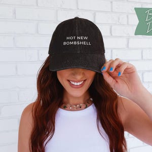 May include: A black denim baseball cap featuring the white embroidered text HOT NEW BOMBSHELL on the front crown. The cap has a structured front panel, a curved brim, and a washed fabric texture. A green tag in the corner displays Black Denim.