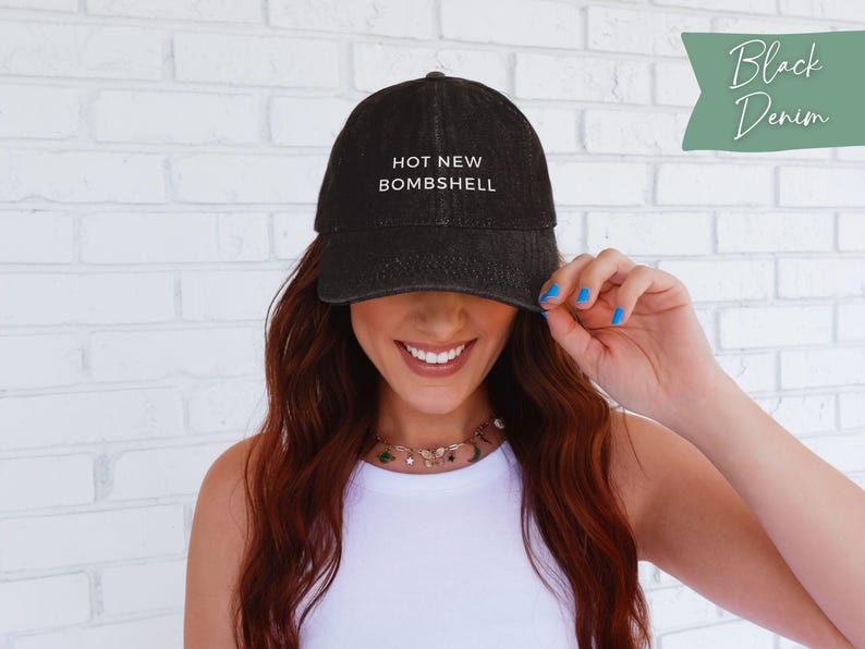 May include: A black denim baseball cap featuring the white embroidered text HOT NEW BOMBSHELL on the front crown. The cap has a structured front panel, a curved brim, and a washed fabric texture. A green tag in the corner displays Black Denim.