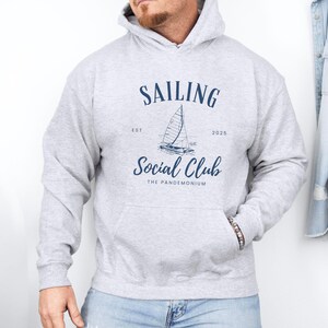 May include: A light gray hoodie with a front pocket and a hood. The navy blue graphic on the front reads "SAILING SOCIAL CLUB" with a sailboat illustration. The text "EST 2020" is also included.