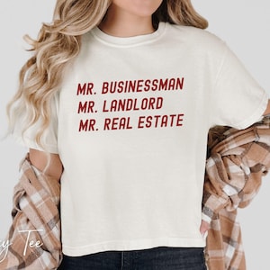 Puede incluir: Camiseta color crema con el texto "MR. BUSINESSMAN, MR. LANDLORD, MR. REAL ESTATE" en rojo. La camiseta se combina con una camisa a cuadros marr&oacute;n y beige. El logotipo de Boxy Tee es visible en la esquina inferior izquierda.
