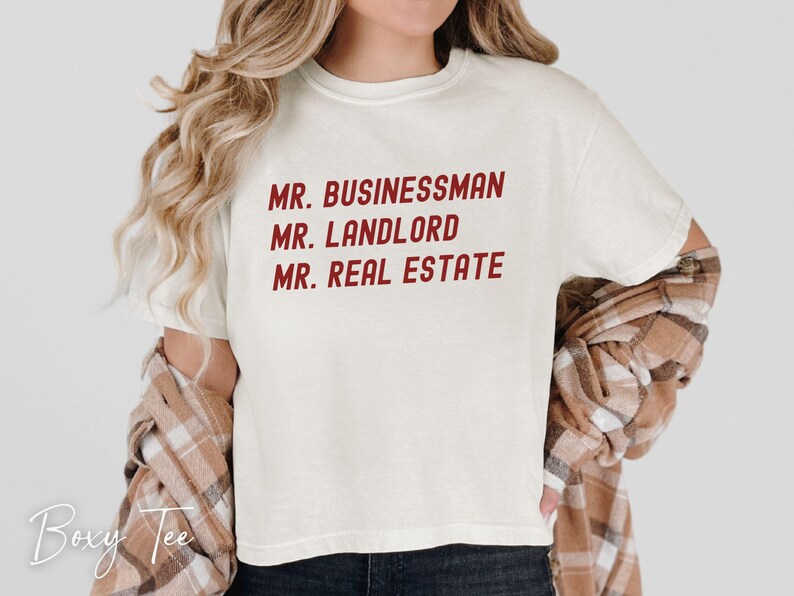 Puede incluir: Camiseta color crema con el texto "MR. BUSINESSMAN, MR. LANDLORD, MR. REAL ESTATE" en rojo. La camiseta se combina con una camisa a cuadros marr&oacute;n y beige. El logotipo de Boxy Tee es visible en la esquina inferior izquierda.