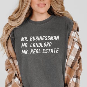 Puede incluir: Camiseta gris oscuro con cuello redondo y las palabras blancas "MR. BUSINESSMAN, MR. LANDLORD, MR. REAL ESTATE". La persona lleva una camisa a cuadros marr&oacute;n y beige sobre la camiseta.