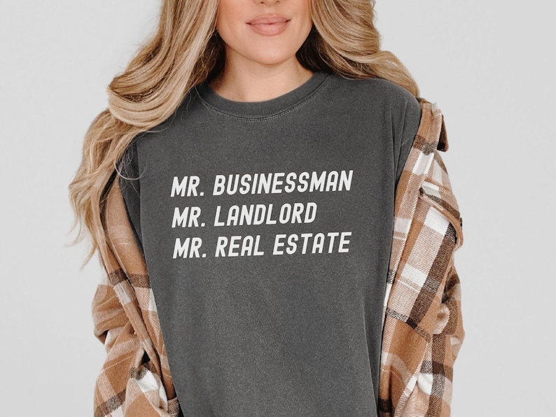 Puede incluir: Camiseta gris oscuro con cuello redondo y las palabras blancas "MR. BUSINESSMAN, MR. LANDLORD, MR. REAL ESTATE". La persona lleva una camisa a cuadros marr&oacute;n y beige sobre la camiseta.
