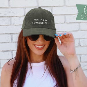 May include: An olive green corduroy baseball cap featuring the text HOT NEW BOMBSHELL embroidered in white on the front panel. The cap has a structured crown, a curved brim, and a button on top, showcasing a textured fabric finish throughout.