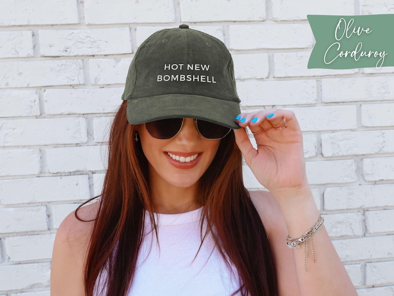 May include: An olive green corduroy baseball cap featuring the text HOT NEW BOMBSHELL embroidered in white on the front panel. The cap has a structured crown, a curved brim, and a button on top, showcasing a textured fabric finish throughout.