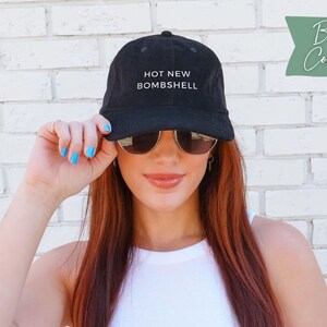 May include: A black corduroy baseball cap featuring the text HOT NEW BOMBSHELL embroidered in white on the front panel. The cap has a structured crown, a curved brim, and a button on top. The fabric has a visible ribbed texture characteristic of corduroy.