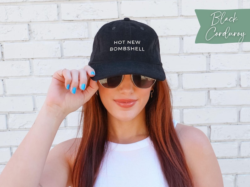 May include: A black corduroy baseball cap featuring the text HOT NEW BOMBSHELL embroidered in white on the front panel. The cap has a structured crown, a curved brim, and a button on top. The fabric has a visible ribbed texture characteristic of corduroy.