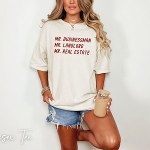 Puede incluir: Camiseta unisex color crema con las palabras "MR. BUSINESSMAN, MR. LANDLORD, MR. REAL ESTATE" en borgo&ntilde;a. La modelo lleva pantalones cortos vaqueros y sostiene un vaso con pajita. Las palabras "Unisex Tee" est&aacute;n escritas en cursiva.