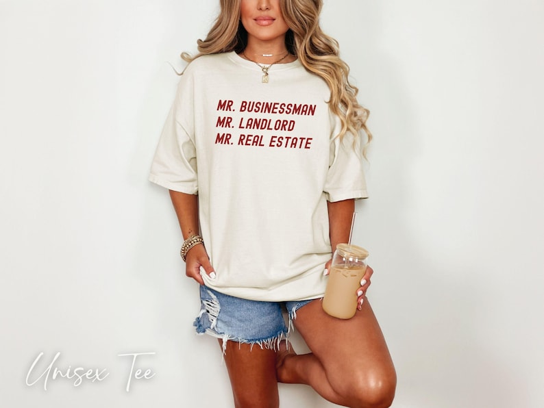 Puede incluir: Camiseta unisex color crema con las palabras "MR. BUSINESSMAN, MR. LANDLORD, MR. REAL ESTATE" en borgo&ntilde;a. La modelo lleva pantalones cortos vaqueros y sostiene un vaso con pajita. Las palabras "Unisex Tee" est&aacute;n escritas en cursiva.