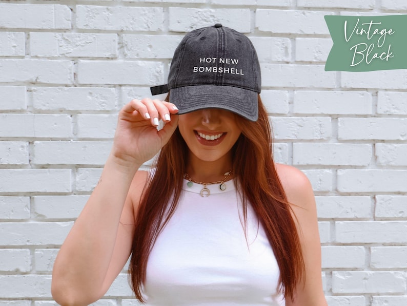 May include: A vintage black baseball cap featuring the white embroidered text HOT NEW BOMBSHELL on the front panel. The cap has a structured crown, a curved brim, and an adjustable strap at the back for a secure fit, showcasing a washed-out aesthetic.