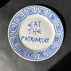 May include: White ceramic plate with a blue geometric border design. The plate features the phrase "EAT THE PATRIARCHY" in blue handwritten text. The plate is round and sits on a dark gray surface.