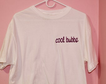 May include: A white t-shirt with the phrase "cool bubbe" in maroon script on the front. The shirt is hanging on a white hanger against a pink wall.